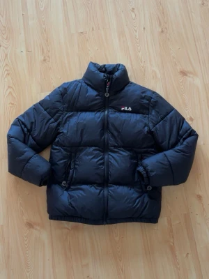 Svart pufferjacka från FILA - Snygg svart pufferjacka från FILA med hög krage och dragkedja framtill. Jackan har två sidofickor med dragkedja och diskret FILA-logga på bröstet. Perfekt för kalla dagar tack vare det vadderade materialet och den klassiska boxiga passformen.