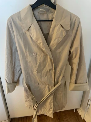 Beige trenchcoat med bälte - Stilren beige trenchcoat med dubbelknäppning och klassisk krage. Jackan har ett matchande bälte med metallspänne i midjan och uppvikta ärmar. Tillverkad i ett lätt, glansigt material som ger en modern look. Perfekt för lager-på-lager-stil.