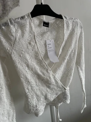 Vit spetsomlottopp från Gina Tricot - Säljer en vit omlottopp från Gina Tricot i spets med långa ärmar och knyt i midjan. Toppen har v-ringning och ett transparent, romantiskt utseende som passar perfekt till jeans eller kjol.