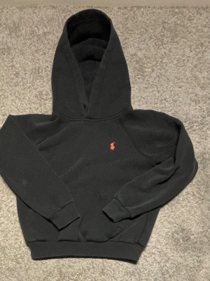 Svart hoodie från Polo Ralph Lauren - Klassisk svart hoodie från Polo Ralph Lauren med röd broderad logga på bröstet. Hoodien har huva, ribbade muddar vid ärmslut och nederkant samt känguruficka framtill. Perfekt för en avslappnad och stilren look.