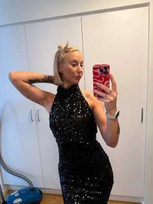 Svart glittrig paljettklänning - Säljer en svart kort paljettklänning med hög hals och öppen rygg. Klänningen är täckt av glittrande paljetter och har en tight passform som framhäver figuren. Perfekt för dig som vill sticka ut med en snygg och festlig look.