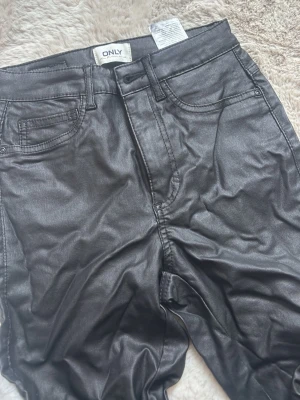 Svarta coated jeans från ONLY - Snygga svarta coated jeans från ONLY med fem fickor och hög midja. Jeansen har en glansig finish som ger en cool läderlook och raka ben. Perfekta för dig som vill ha en trendig och edgy stil.