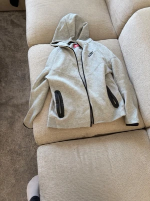 Grå hoodie från Nike med dragkedja - Snygg grå hoodie från Nike med svart logga på bröstet och hel dragkedja framtill. Två fickor med svarta detaljer och huva. Tillverkad i mjukt sweatshirtmaterial som är perfekt för chill eller träning.