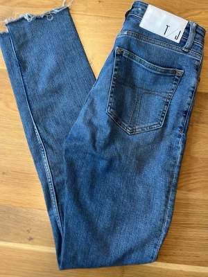 Blå raka jeans från T/J - Säljer ett par klassiska blå jeans från T/J med raka ben och råa, fransiga benslut. Jeansen har en bakficka med dekorativ söm och en vit patch med logga i midjan. Perfekta till en avslappnad och trendig look.