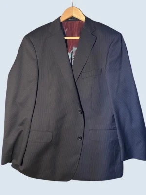 Oscar Jacobson Mörkgrå kavaj med diskreta ränder - Stilren mörkgrå kavaj från Oscar Jacobson med tunna, diskreta ränder. Klassisk tvåknäppt modell med tre fickor framtill och en innerficka. Fodret är vinrött och glansigt, vilket ger en snygg kontrast. Perfekt för en dressad look.