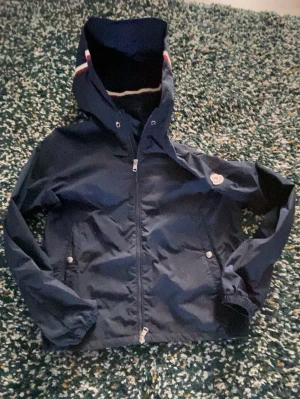 Mörkblå vindjacka från Moncler - Snygg mörkblå vindjacka från Moncler med huva och dragkedja framtill. Jackan har vita och röda detaljer på huvan, två fickor med tryckknappar och elastiska muddar vid ärmsluten. Tillverkad i ett lätt och vattenavvisande material, perfekt för blåsiga dagar.
