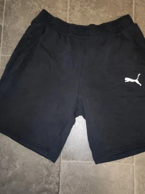 Svarta shorts från Puma - Svarta shorts från Puma med elastisk midja och två sidofickor. Klassisk Puma-logga i vitt på vänster ben. Perfekta för träning eller chill, tillverkade i mjukt bomullsmaterial för skön känsla.