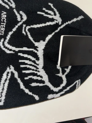 Svart mössa från Arc'teryx - Snygg svart mössa från Arc'teryx med vitt grafiskt skelettmönster och broderad logga på sidan. Tillverkad i mjukt stickat material som sitter skönt på huvudet. Perfekt för dig som gillar stilrena accessoarer med en sportig touch.