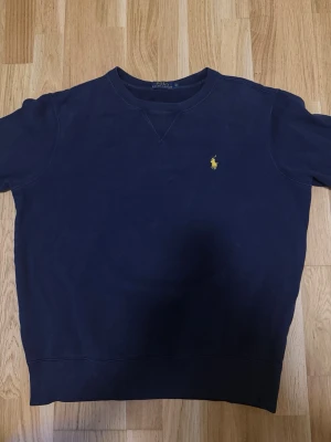 Mörkblå sweatshirt från Polo Ralph Lauren - Klassisk mörkblå sweatshirt från Polo Ralph Lauren med gul broderad logga på bröstet. Tröjan har rund halsringning och ribbade muddar vid ärmslut och nederkant. Perfekt för en avslappnad och stilren look.