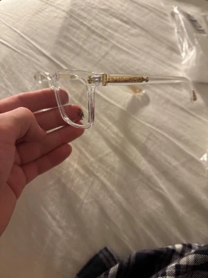 Chrome heart glasögon - Säljer ett par stilrena glasögon med transparent båge och rektangulär form. Skalmarna har snygga guldfärgade detaljer och ett litet svart korsmönster på ena linsen. Perfekt accessoar för dig som vill sticka ut med en unik look.