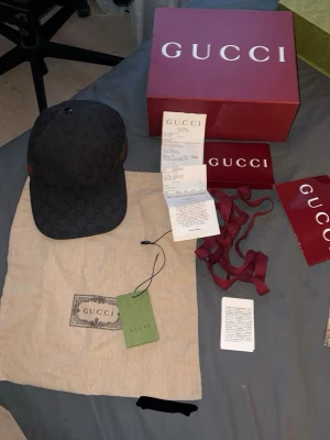 Svart keps från Gucci med monogram - Säljer en svart keps från Gucci med det klassiska GG-monogrammönstret. Kepsen har böjd skärm och diskreta gröna och röda detaljer på sidorna. Tillverkad i canvasmaterial och kommer med originalkartong, dustbag och tags.