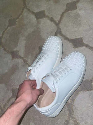 Vita sneakers med nitar från Louboutin - Säljer ett par vita sneakers från Christian Louboutin med coola nitar på tån. Skorna är i slätt läder och har vita skosnören samt en klassisk låg siluett. Insidan är beige och sulan är vit. Perfekta för dig som vill sticka ut med din stil.