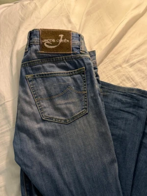 Jacob Cohen jeans - Schyssta blå jeans från Jacob Cohen med klassisk femficksdesign och ljus tvätt. Jeansen har raka ben och detaljerade sömmar, samt en brun patch med logga bak i midjan. Perfekta för en avslappnad och trendig stil. Lite slitningar gjorda själv men syns knappt när man har på dom
