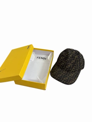 Fendi keps med FF-mönster - Snygg keps från Fendi i brunt och svart med det klassiska FF-logomönstret över hela kepsen. Kepsen har böjd skärm och är tillverkad i ett tygmaterial som känns lyxigt och trendigt. Perfekt accessoar för att lyfta din streetstyle.