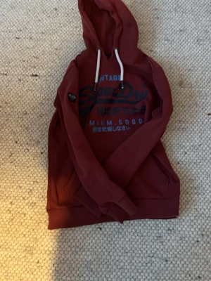Vinröd hoodie från Superdry - Snygg vinröd hoodie från Superdry med tryck i blått och vitt på bröstet samt japanska tecken. Hoodien har vita dragsnören, känguruficka och ribbade muddar. Perfekt för en avslappnad och trendig stil.
