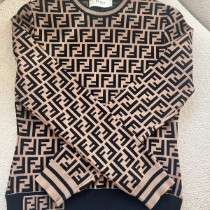 Beige och svart mönstrad tröja från Fendi - Snygg beige och svart tröja från Fendi med ett ikoniskt mönster. Tröjan har långa ärmar och ribbade muddar vid ärmslut och nederkant. Perfekt för att ge din outfit en lyxig touch. Strl M men passar som en S (pris kan diskuteras)