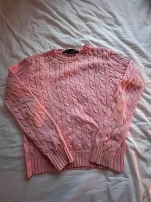 Rosa kabelstickad tröja från Ralph Lauren - Snygg kabelstickad tröja i ljusrosa från Ralph Lauren med klassisk blå logga broderad på bröstet. Tröjan har rund halsringning, långa ärmar och ribbade muddar vid ärmslut och nederkant. Perfekt för lager på lager och en tidlös favorit i garderoben.