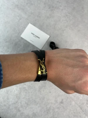 Svart läderarmband från Saint Laurent - Stilrent svart armband i läder från Saint Laurent med dubbla remmar och ikoniskt YSL-spänne i guldmetall. Perfekt accessoar för att lyfta din outfit med en lyxig touch och klassisk design.
