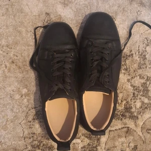 Christian Louboutins  - Snygga svarta sneakers från Christian Louboutin med klassisk röd sula. Skorna har rund tå, snörning och är tillverkade i mocka för en lyxig känsla. Perfekta för dig som vill sticka ut med en stilren men ändå unik detalj.