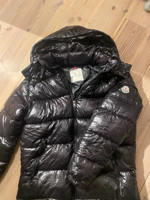 Svart dunjacka från Moncler med luva - Snygg svart dunjacka från Moncler med glansig finish och avtagbar luva. Jackan har dragkedja framtill, två sidofickor med dragkedja och Moncler-logga på ärmen. Perfekt för kalla vinterdagar och riktigt varm tack vare dunfyllningen. Tveka inte vid funderingar!
