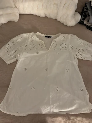 Vit blus med broderade detaljer - Söt vit blus från AMELIE & ME i storlek S. Blusen har broderade blommönster, korta puffärmar och en v-ringad hals med spetskant. Tillverkad i lätt bomull som känns luftig och perfekt för varmare dagar.