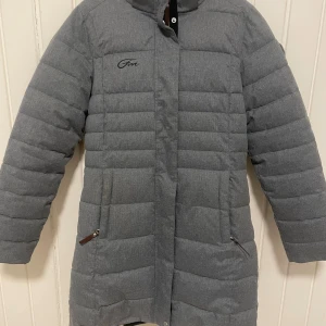 Grå dunjacka med fuskpäls från Five Seasons  - Snygg grå dunjacka från Give med avtagbar huva och fuskpäls. Jackan har två dragkedjefickor, diskret logga på bröstet och quiltad design. Perfekt för kalla vinterdagar och riktigt varm tack vare dunfyllningen.