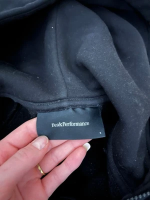Svart hoodie Peak Performance S - Svart hoodie från Peak Performance med dragkedja framtill och två stora fickor. Klassisk logga broderad på bröstet och justerbar huva med snören. Tillverkad i mjukt bomullsmaterial som känns skönt mot huden. 