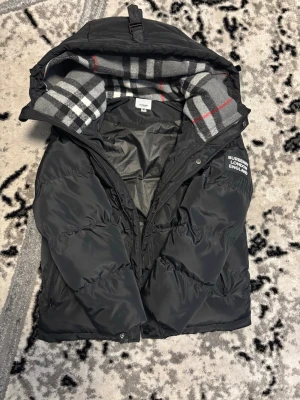 Svart pufferjacka från Burberry - Svart pufferjacka från Burberry med klassiskt rutigt foder i grått, vitt, svart och rött. Jackan har luva, dragkedja och tryckknappar samt en tydlig Burberry London England-logga på sidan av armen. Perfekt för kalla dagar med sin vadderade design och snygga detaljer.