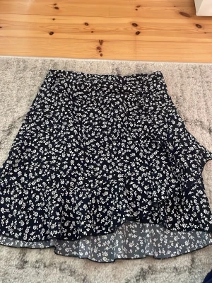 Blommig volangkjol i mörkblått - Supersöt kort kjol med volangkant och blommigt mönster i vitt på mörkblå botten. Kjolen har omlottkänsla och är perfekt för dig som gillar en feminin och trendig stil. Lätt och luftig, passar perfekt till varmare dagar.