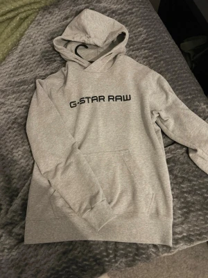 Grå hoodie från G-Star RAW - Säljer en stilren grå hoodie från G-Star RAW med svart logga tryckt på bröstet. Hoodien har en klassisk känguruficka framtill och ribbade muddar vid ärmslut och nederkant. Perfekt för en avslappnad och trendig look.