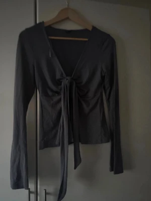 Mörkgrå långärmad topp med knyt - Trendig mörkgrå långärmad topp med djup v-ringning och knyt framtill. Figurnära passform och mjukt stretchigt material som ger en snygg siluett. Perfekt att styla med jeans eller kjol för en cool look. För fler bilder/information skriv gärna, priset kan justeras 🤗❤️
