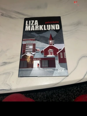 Polcirkeln av Liza Marklund - Säljer boken 'Polcirkeln' av Liza Marklund, utgiven av Piratförlaget. Omslaget har en illustration i grå, röd och vit färg med motiv av ett bibliotek och en kyrka i norrländsk miljö. Perfekt för dig som gillar spännande skönlitteratur och deckare.