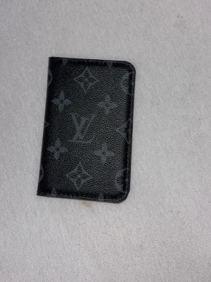 Svart plånbok - Snygg svart plånbok med klassiskt monogrammönster i grått. Plånboken är i läder och har flera kortfack på insidan. Perfekt för dig som vill ha en stilren och lyxig accessoar i fickan.