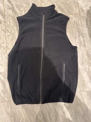 Svart fleeceväst med dragkedja - En stilren svart fleeceväst med hög krage och hel dragkedja framtill. Västen har två fickor med dragkedja på sidorna och är ärmlös för en avslappnad look. Perfekt att slänga på över en hoodie eller tröja för extra värme.