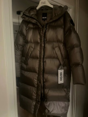 Lång brun dunjacka från 8848 Altitude - Snygg lång brun dunjacka från 8848 Altitude med huva och dragkedja framtill. Jackan har en puffig design, stora paneler och sidofickor. Perfekt för kalla vinterdagar och ger en riktigt trendig look.