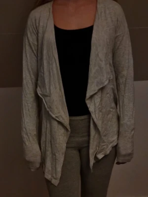 Ljusgrå öppen kofta - Säljer en ljusgrå, långärmad öppen kofta med avslappnad passform och draperad framsida. Koftan är tillverkad i ett mjukt och tunt material, perfekt att slänga över en topp för en chill look.