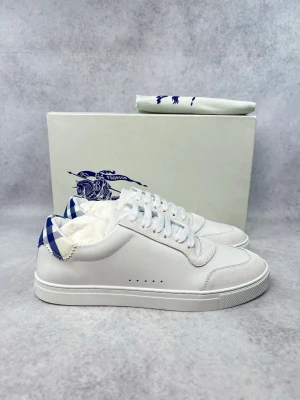 Vita sneakers från Burberry - Stilrena vita sneakers från Burberry med blåvitt rutigt mönster på hälen. Skorna har ovandel i läder och mockadetaljer framtill, samt vita skosnören och diskreta perforeringar på sidan. Klassisk låg modell som passar till det mesta.