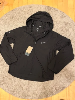 Svart Nike vindjacka med huva - Snygg svart vindjacka från Nike med huva och dragkedja framtill. Jackan har reflekterande detaljer, två sidofickor och Nike-logga på bröstet. Tillverkad i lätt och slitstarkt material, perfekt för löpning eller promenader. Justerbar huva och sportig passform.