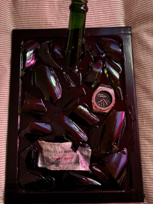 Unik klocka i konstverk med glas - Säljer en unik klocka infälld i ett konstverk av mörkgröna glasbitar och en flaskhals på svart platta. Klockan har rund urtavla med svart bakgrund och vita detaljer. Perfekt för dig som gillar annorlunda accessoarer och konstnärlig stil.