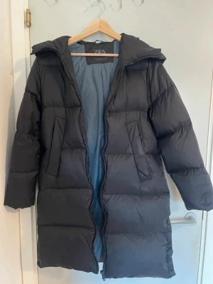 Svart lång dunjacka från Zara - Svart lång dunjacka från Zara med huva och dragkedja framtill. Jackan har två snedställda fickor med lock och quiltad design. Perfekt för kalla vinterdagar och har en stilren look som passar till det mesta. Passar mig som bär XS/S