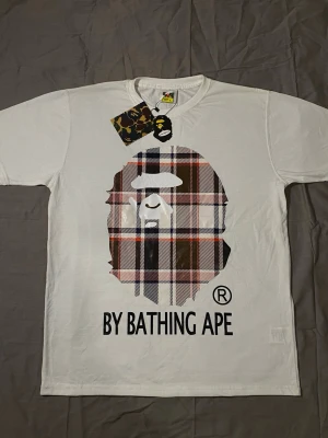 BAPE X Burberry - Vit t-shirt från BAPE med klassisk passform och korta ärmar. Stor rutig BAPE-logga framtill i brunt, svart, vitt och rött samt texten 'By Bathing Ape' under. Tillverkad i mjuk bomull och perfekt för dig som gillar streetwear.