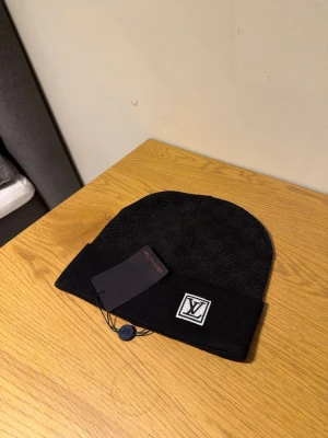 Svart mössa från Louis Vuitton - Snygg svart mössa från Louis Vuitton med uppvikt kant och klassisk LV-logga framtill. Tillverkad i mjukt stickat material som håller dig varm och stilren under kyliga dagar. Perfekt accessoar för dig som vill ha en clean och exklusiv look.