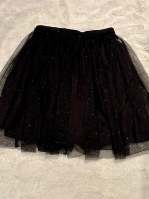 Svart glittrig tyllkjol - Säljer en svart kortkjol i tyll med glittriga detaljer. Kjolen har flera lager för extra volym och en elastisk midja som gör den bekväm att bära. Perfekt för dig som vill sticka ut lite extra med en glittrig look.