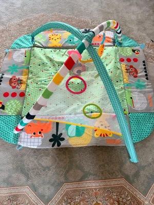 Färgglad babygym  - Säljer ett babygym i tyg med färgglada djurmotiv som giraff, apa och elefant. Gymmet har två bågar i olika färger, mjuk matta i grönt med prickar och flera ringar för leksaker. Kan användas både som lekmatta och med uppfällda kanter.