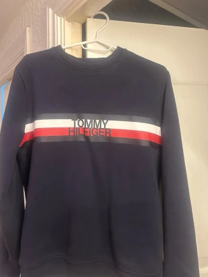 Mörkblå sweatshirt Tommy Hilfiger - Snygg mörkblå sweatshirt från Tommy Hilfiger med klassiska ränder i vitt och rött över bröstet samt logga framtill. Tröjan har rund halsringning och långa ärmar. Perfekt för en avslappnad och stilren look.