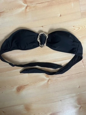 Svart bandeau bikiniöverdel med gulddetalj - Snygg svart bandeau bikiniöverdel med en unik guldfärgad ring i mitten. Knytband baktill för justerbar passform. Perfekt för stranden eller poolen och ger en stilren look.