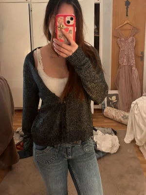 Glittrig svart kofta från Vero Moda - Säljer en svart glittrig kofta från Vero Moda med v-ringning och knappar framtill. Koftan har långa ärmar och ribbade muddar vid ärmslut och nederkant. Perfekt att slänga över ett linne eller t-shirt för en snygg layering-effekt.