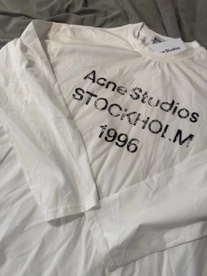 Vit långärmad t-shirt Acne Studios - Vit långärmad t-shirt från Acne Studios med svart tryck på bröstet där det står 'Acne Studios STOCKHOLM 1996'. Klassisk passform och rund halsringning. Perfekt för en stilren och avslappnad look. Storlek L
