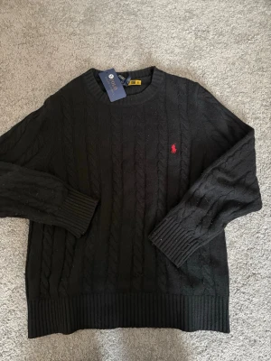 Svart kabelstickad tröja Polo Ralph Lauren - Svart kabelstickad tröja från Polo Ralph Lauren med klassisk rund halsringning och röd broderad logga på bröstet. Tröjan har ribbade muddar och nederkant, perfekt för en stilren look. Den är knappt använd och passar dig mellan 165 - 180!