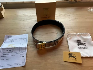 Randigt Burberry bälte med guldspänne - 105 cm, Säljer ett klassiskt Burberry bälte med det ikoniska rutiga mönstret i vitt, svart och rött. Bältet har ett stort guldigt B-spänne och är Perfekt accessoar för att lyfta vilken outfit som helst.